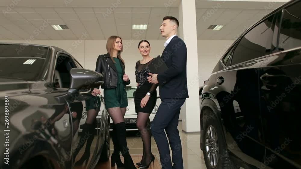 Two gorgeous girls talking to a sales consultant in a car dealership, they want to buy a new car. The process of working in the showroom. Two gorgeous girls talking to a sales consultant in a car dealership, they want to buy a new car. The process of working in the showroom.