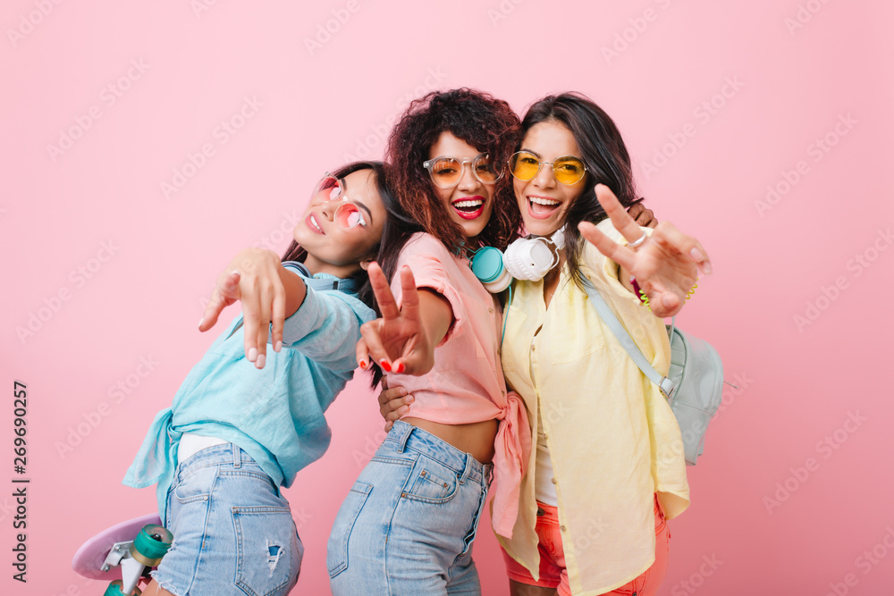 Hispanic funny girl pointing with finger while posing with best friends. Sporty female model in headphones joking with asian ladies. Hispanic funny girl pointing with finger while posing with best friends. Sporty female model in headphones joking with asian ladies.
