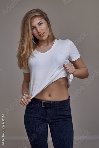 Attractive slender sexy young blond woman in jeans and a white T-shirt lifting the corner of her top to expose a section of her abdomen while giving the camera a sultry look with parted lips
