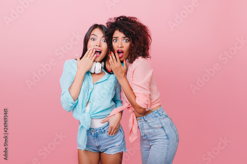 Sensual caucasian woman with black short hair posing with shocked face expression on pink background. Surprised dark-haired girl embracing latin female friend.