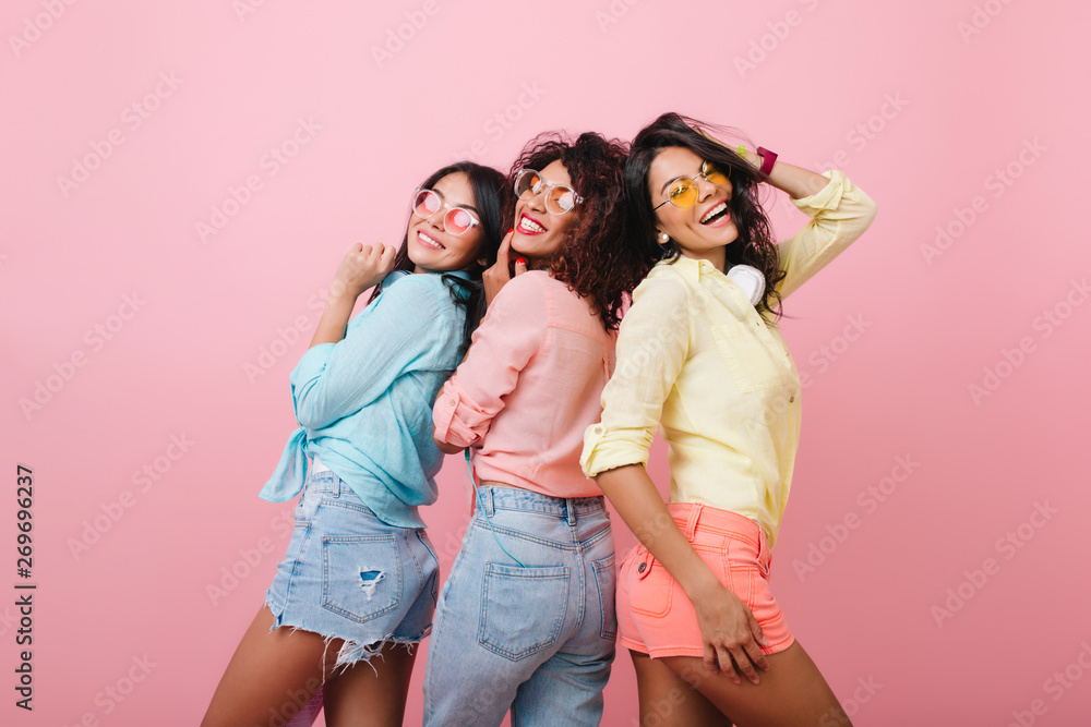 Carefree girls in colorful cotton shirts posing together and smiling. Indoor portrait of attractive young ladies expressing happy emotions during joint photoshoot. - obrazy, fototapety, plakaty 