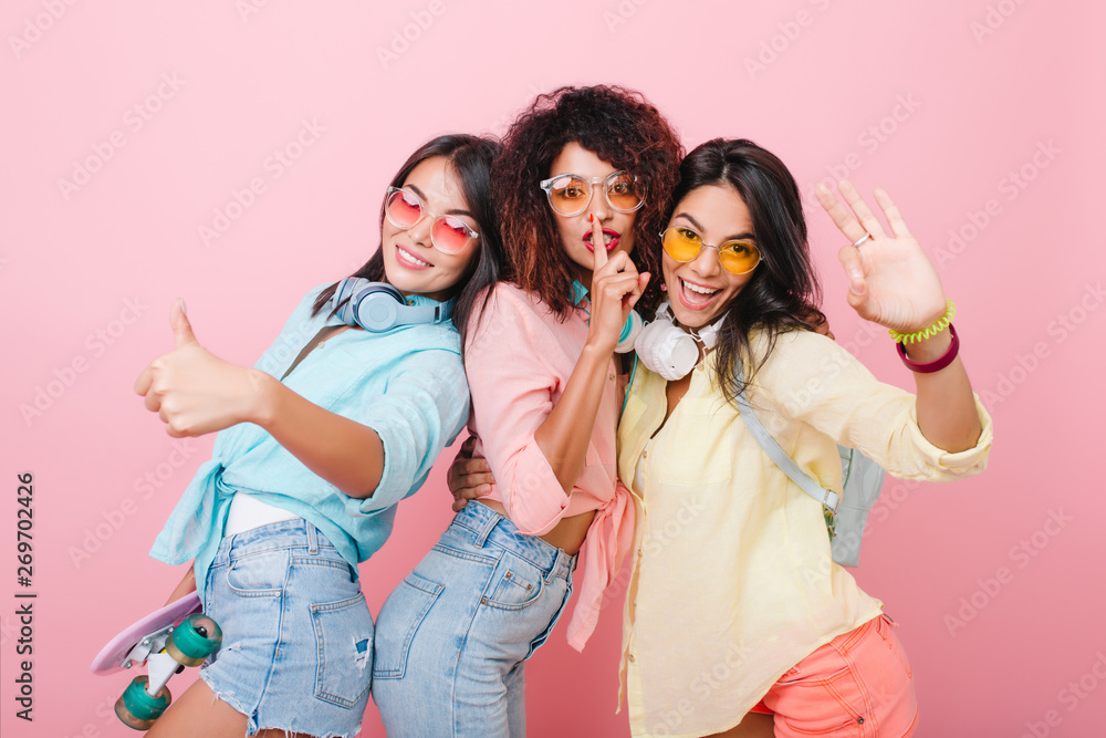 Refined hispanic woman in colorful bracelets waving hand during joint photoshoot with friends. Lovely asian lady in stylish blue shirt laughing while spending time with girls. Refined hispanic woman in colorful bracelets waving hand during joint photoshoot with friends. Lovely asian lady in stylish blue shirt laughing while spending time with girls.