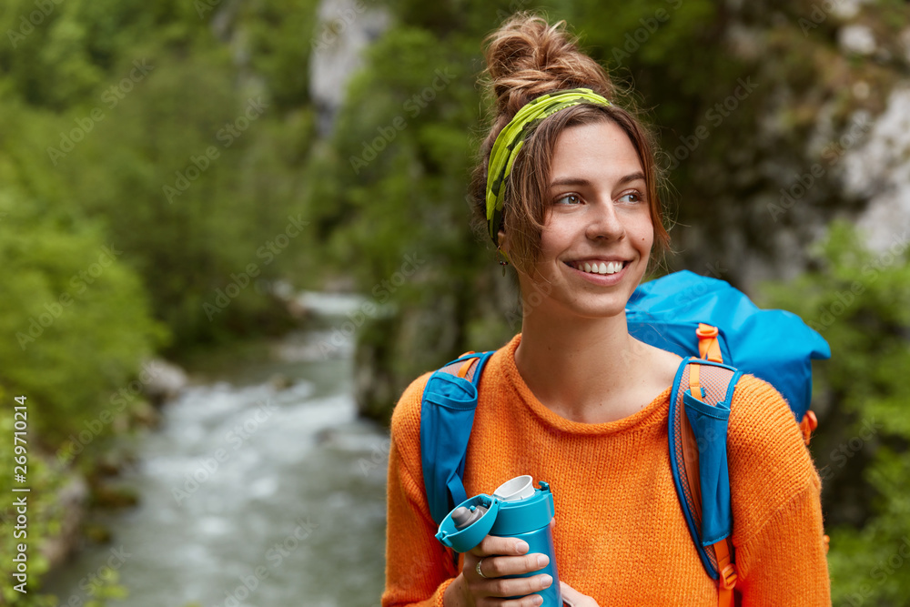 Φωτογραφία Glad tourist recreats in mountains near small river, green trees, focused aside with broad smile, dressed in casual clothes, has big rucksack on shoulders, drinks aromatic bevergae from flask
