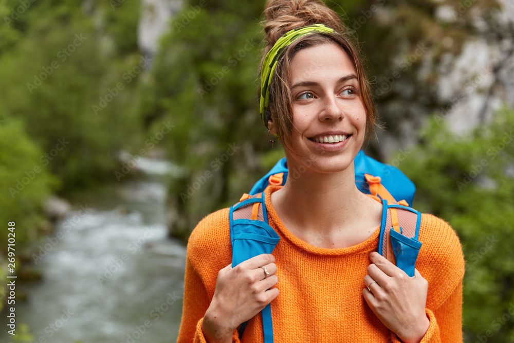 Φωτογραφία Pensive glad woman travels in majestic place, looks happily away, wears casual orange sweater, carries rucksack, spends nice time in green mountain forest, poses over stream