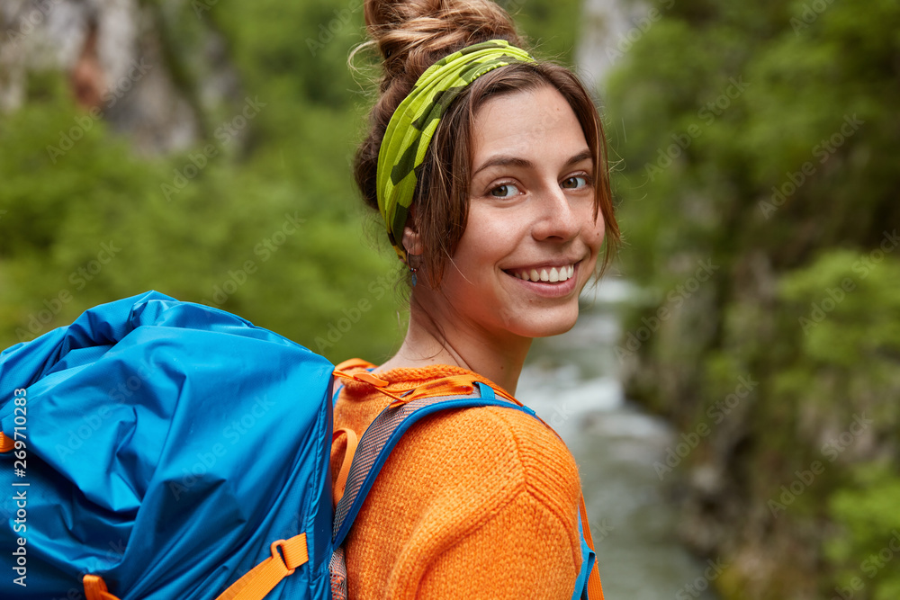 Φωτογραφία Happy girl traveler has exploration trip, wears rucksack, stands beside stream, smiles broadly, has pleasant appearance, blank space against nature background, feels joyful