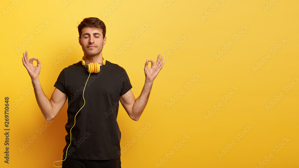 Concentrated young European man keeps hands in okay gesture, meditates indoor, has eyes closed, tries to relax with calm music in headphones, dressed in casual outfit, isolated on yellow wall Concentrated young European man keeps hands in okay gesture, meditates indoor, has eyes closed, tries to relax with calm music in headphones, dressed in casual outfit, isolated on yellow wall