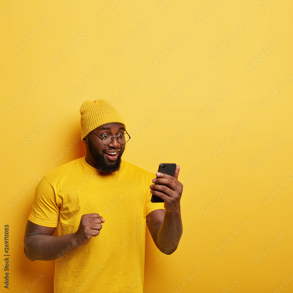 Overjoyed Afro man with thick bristle, looks happily at cell phone, feels upbeat, celebrates good news, keeps clenched fist, wears yellow stylish hat, t shirt, models in studio, blank space upwards Overjoyed Afro man with thick bristle, looks happily at cell phone, feels upbeat, celebrates good news, keeps clenched fist, wears yellow stylish hat, t shirt, models in studio, blank space upwards