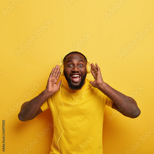 Joyous dark skinned guy has fun indoor, enjoys melomany time, holds hands on headphones, enjoys good sound as listens favourite audio track, wears yellow t shirt, stands indoor. People and music © Wayhome Studio