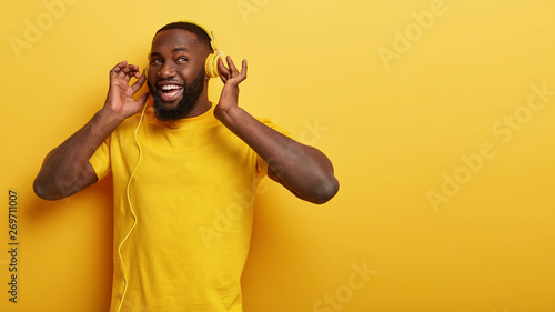 Technology and lifestyle concept. Positive unshaven guy listens favourite music, enjoys great song, uses modern headphones connected to some device, wears casual clothes, isolated over yellow wall
