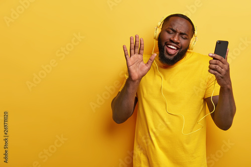 Overjoyed Afro American guy sings song while listens music in headphones, dances and moves actively, smiles broadly, dressed in casual t shirt, isolated over yellow background with copy space