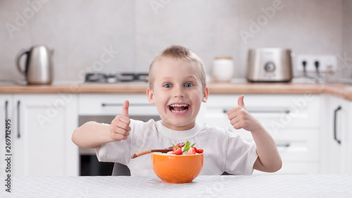 Little caucasian smiling boy sitting at the kitchen table and eat porridge with strawberry