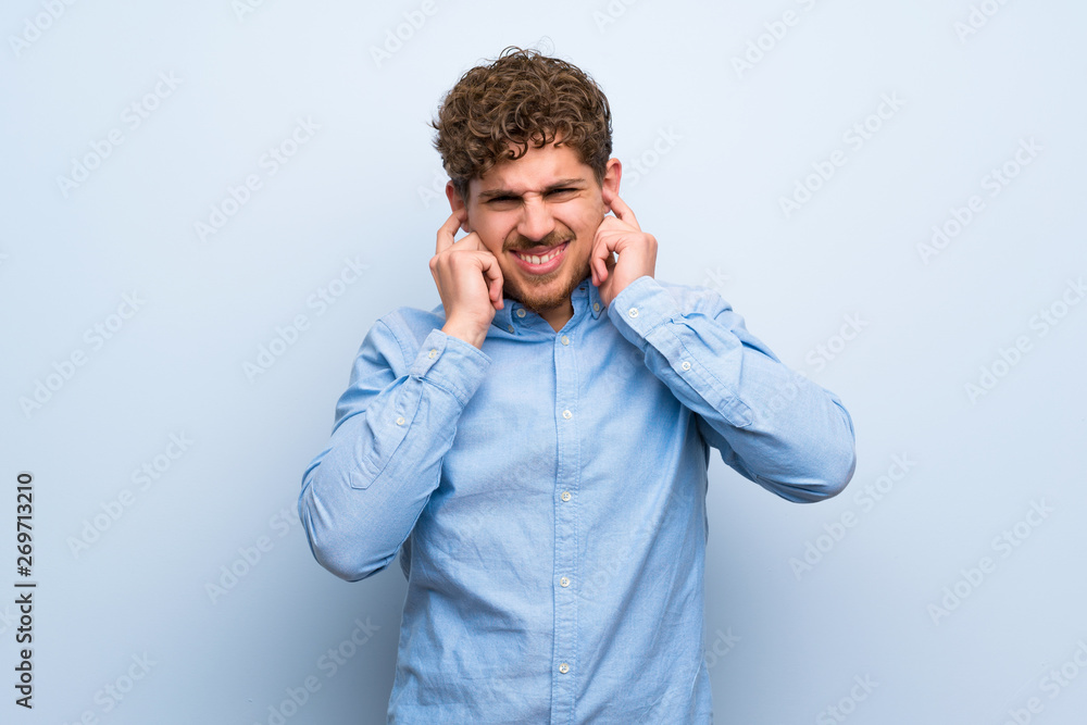 Blonde man over blue wall frustrated and covering ears Blonde man over blue wall frustrated and covering ears