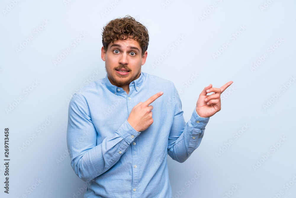 Blonde man over blue wall frightened and pointing to the side Blonde man over blue wall frightened and pointing to the side