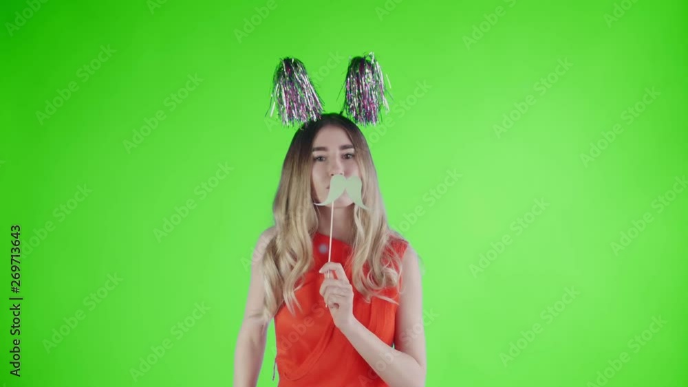 Beautiful girl in party hat and cardboard mustache is dancing on a green screen . Beautiful girl in party hat and cardboard mustache is dancing on a green screen .