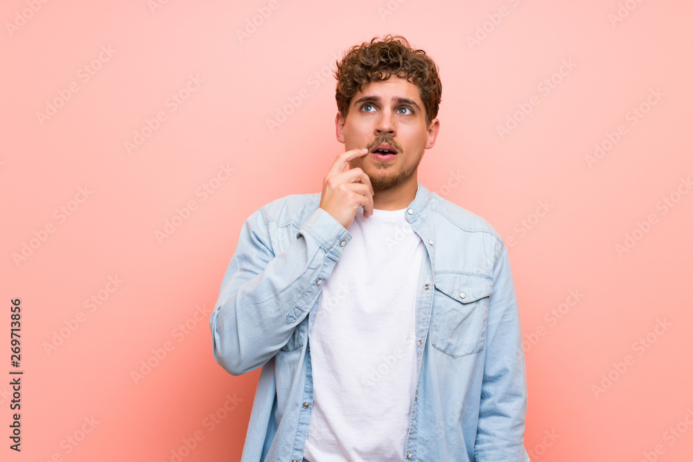 Blonde man over pink wall having doubts while looking up Blonde man over pink wall having doubts while looking up