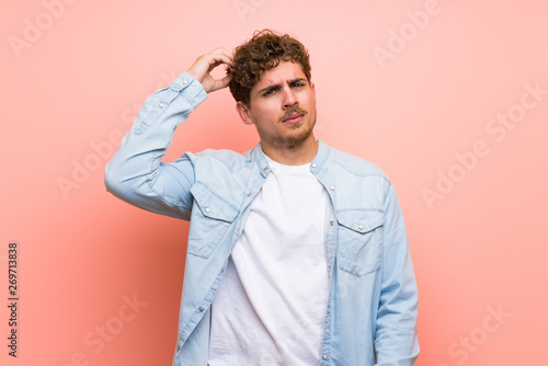 Blonde man over pink wall having doubts while scratching head