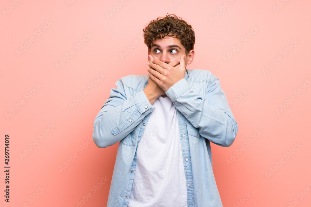 Blonde man over pink wall covering mouth with hands