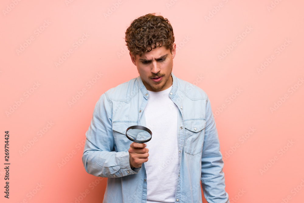 Blonde man over pink wall holding a magnifying glass Blonde man over pink wall holding a magnifying glass