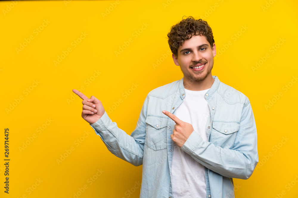 Blonde man over isolated yellow wall pointing finger to the side Blonde man over isolated yellow wall pointing finger to the side