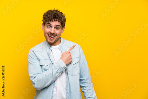 Blonde man over isolated yellow wall pointing finger to the side