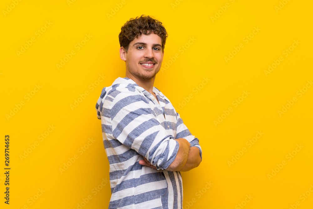Blonde man over yellow wall keeping the arms crossed in lateral position while smiling Blonde man over yellow wall keeping the arms crossed in lateral position while smiling