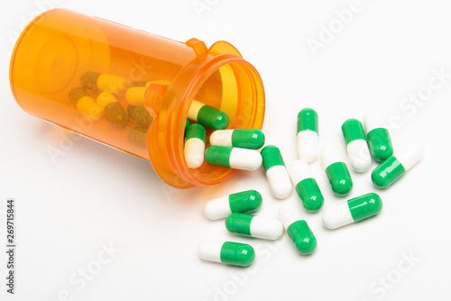 A bunch of green and white capsules spilling out of a slightly transparent orange medicine pharmacy bottle set on a plain white background in horizontal image format.