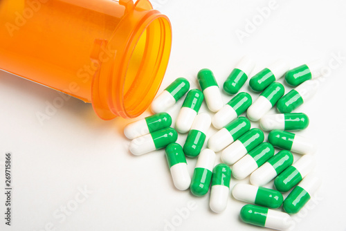 An open and capless orange plastic medicine bottle with white and green pills spilling out and into a plain white background.