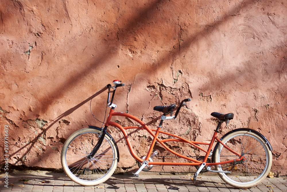 Modern comfortable orange tandem double bicycle with white wheels on paved empty sidewalk leaned against old plastered cracked red wall on bright sunny summer day. Transport and traveling concept. Modern comfortable orange tandem double bicycle with white wheels on paved empty sidewalk leaned against old plastered cracked red wall on bright sunny summer day. Transport and traveling concept.