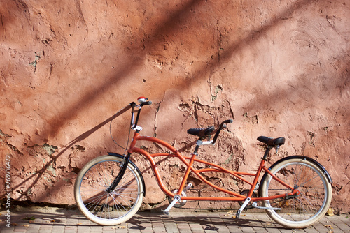 Modern comfortable orange tandem double bicycle with white wheels on paved empty sidewalk leaned against old plastered cracked red wall on bright sunny summer day. Transport and traveling concept.