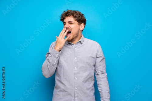 Blonde man over blue wall yawning and covering wide open mouth with hand