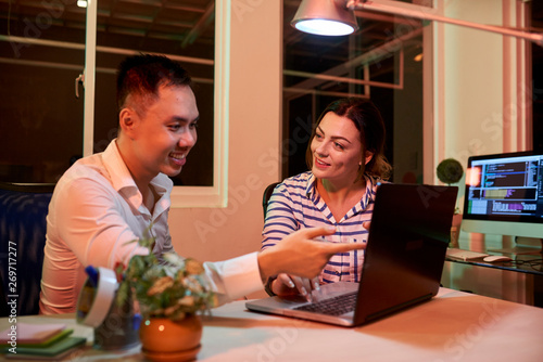 Smiling Asian businessman pointing at monitor of laptop computer and talking with businesswoman listening to him, they working at office till night