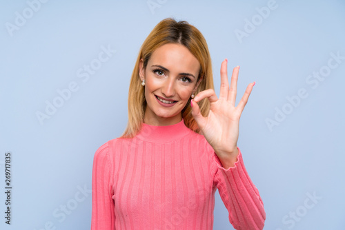 Young blonde woman with pink sweater over isolated blue background showing an ok sign with fingers