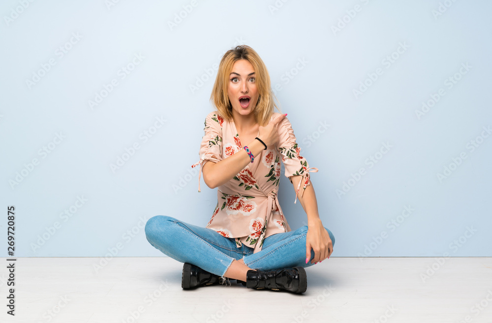Young blonde woman sitting on the floor surprised and pointing side