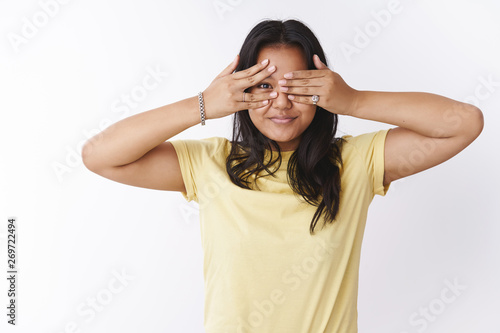 Young polynesian girl cover eyes and peeking through fingers smiling broadly anticiapting amazing surprise standing lovely and cute against white background in casual yellow t-shirt