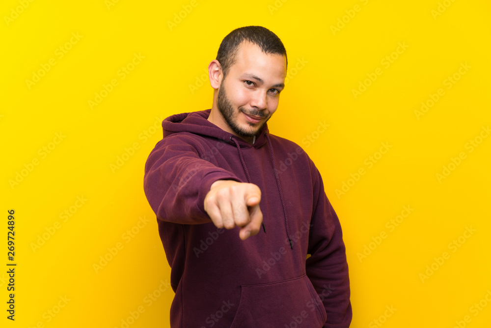 Colombian man with sweatshirt over yellow wall points finger at you with a confident expression Colombian man with sweatshirt over yellow wall points finger at you with a confident expression