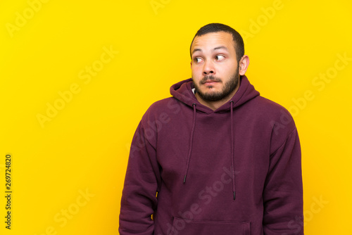Colombian man with sweatshirt over yellow wall making doubts gesture looking side