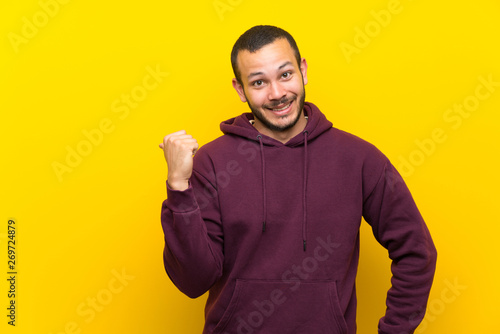 Colombian man with sweatshirt over yellow wall pointing to the side to present a product