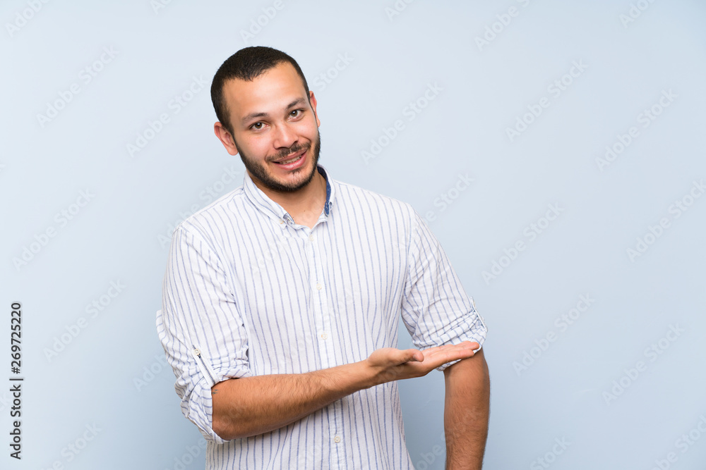 Colombian man over isolated blue wall presenting an idea while looking smiling towards Colombian man over isolated blue wall presenting an idea while looking smiling towards