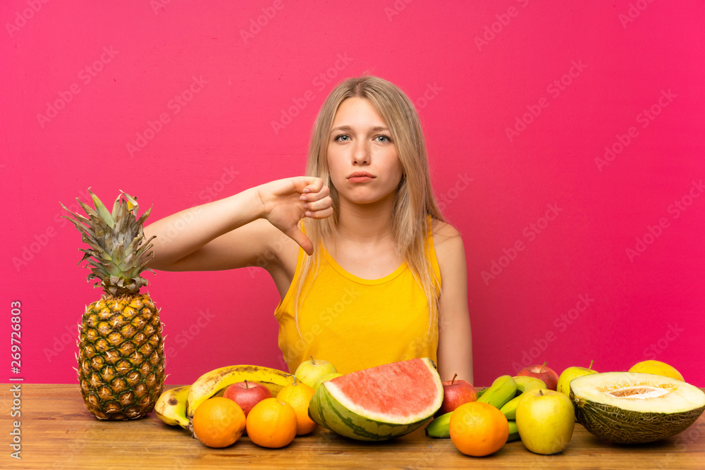 Young blonde woman with lots of fruits showing thumb down sign Young blonde woman with lots of fruits showing thumb down sign