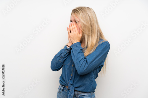Young blonde woman over isolated white wall covering mouth and looking to the side