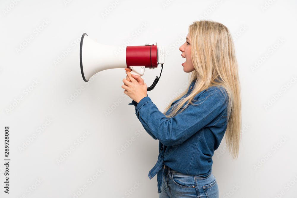 Young blonde woman over isolated white wall shouting through a megaphone Young blonde woman over isolated white wall shouting through a megaphone