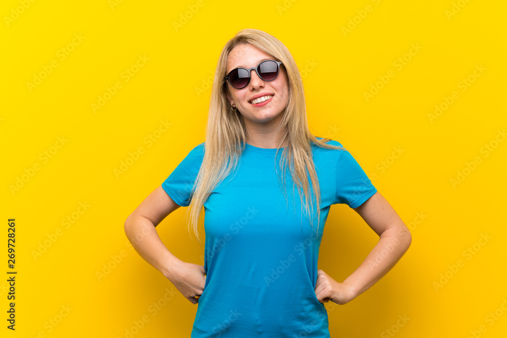 Young blonde woman over isolated yellow background with glasses and smiling Young blonde woman over isolated yellow background with glasses and smiling