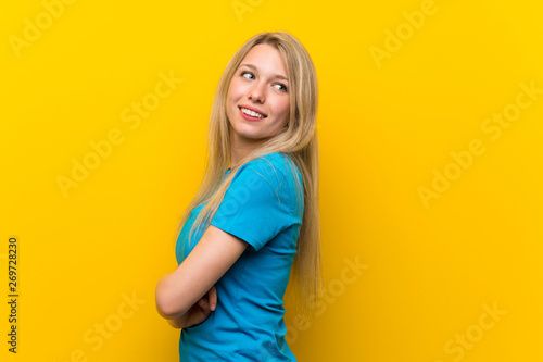 Young blonde woman over isolated yellow background with arms crossed and happy