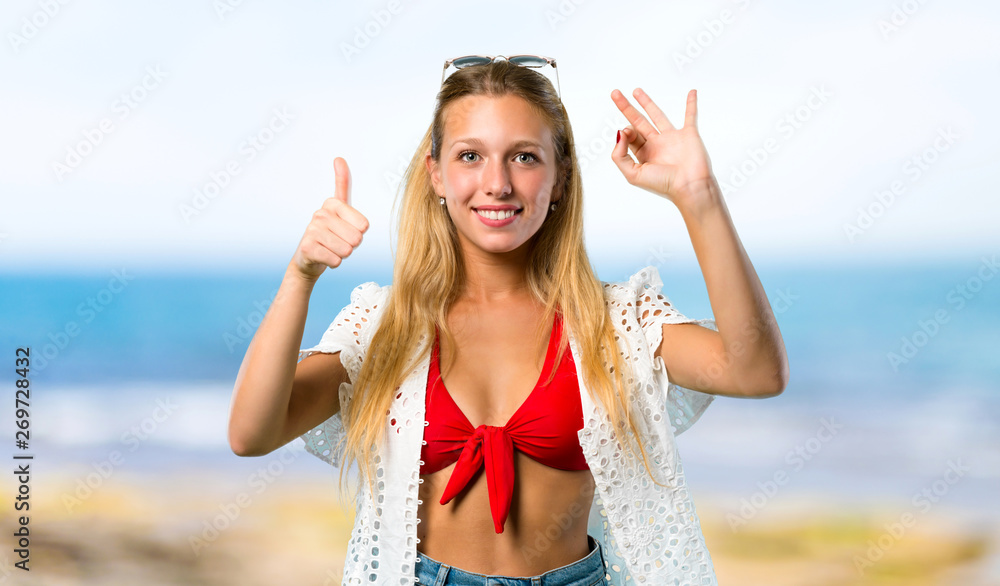 Blonde girl in summer vacation showing ok sign and giving a thumb up gesture with the other hand at the beach