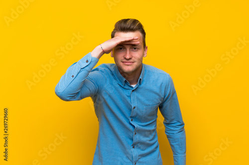 Blonde man over isolated yellow wall looking far away with hand to look something © luismolinero