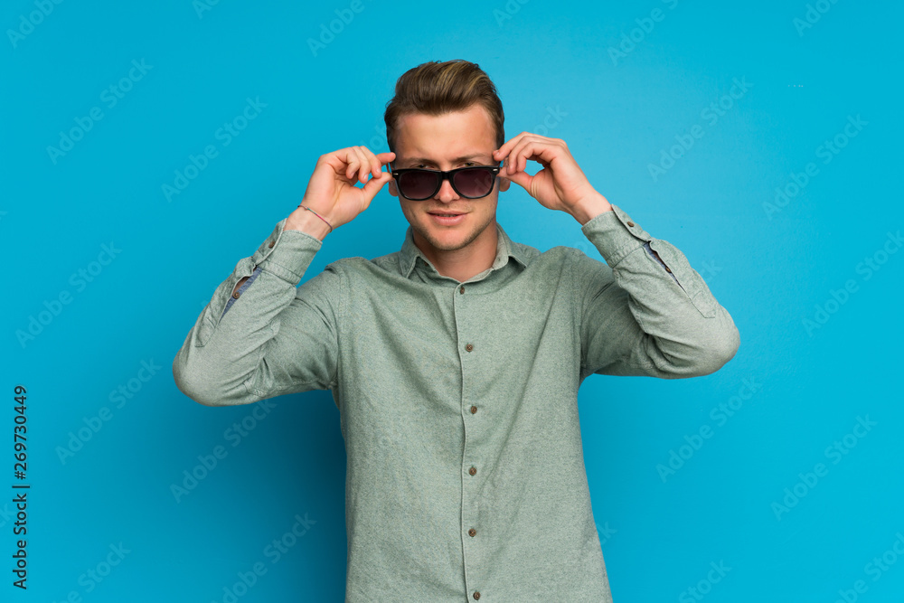 Blonde man over isolated blue wall with glasses and surprised Blonde man over isolated blue wall with glasses and surprised