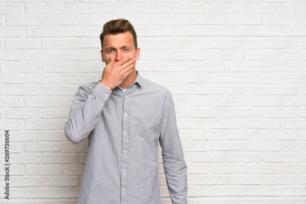 Blonde man over white brick wall covering mouth with hands Blonde man over white brick wall covering mouth with hands
