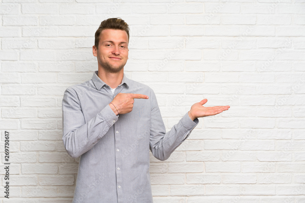 Blonde man over white brick wall holding copyspace imaginary on the palm to insert an ad Blonde man over white brick wall holding copyspace imaginary on the palm to insert an ad