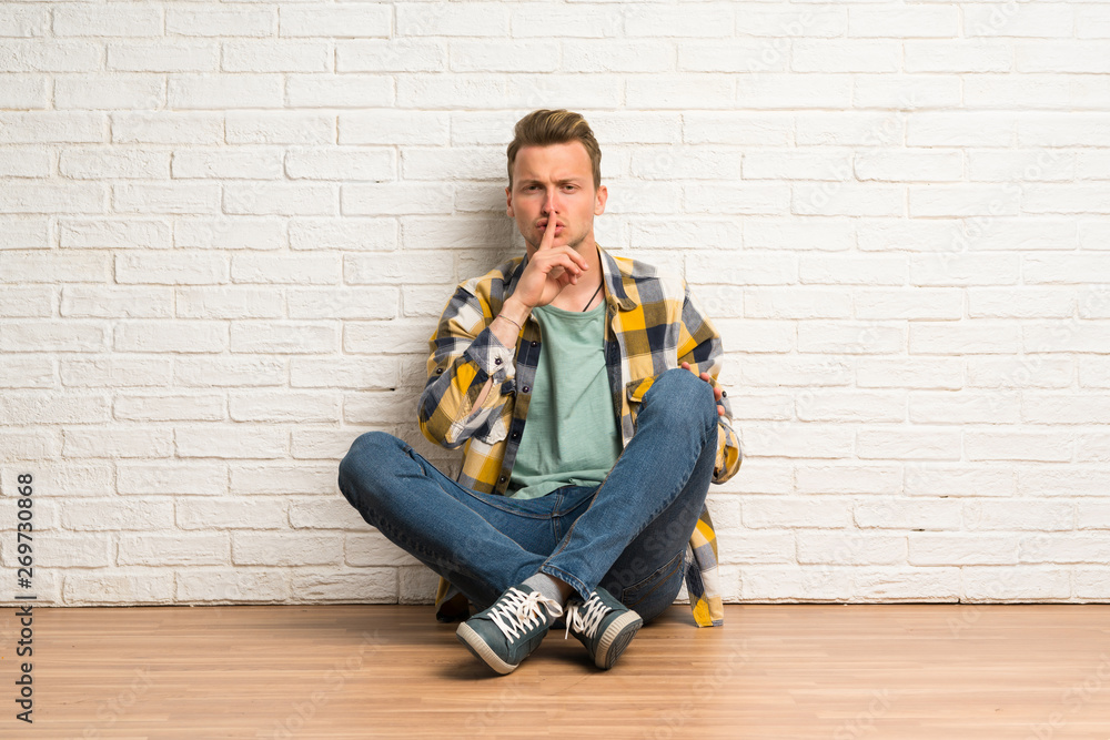 Blonde man sitting on the floor showing a sign of silence gesture putting finger in mouth Blonde man sitting on the floor showing a sign of silence gesture putting finger in mouth
