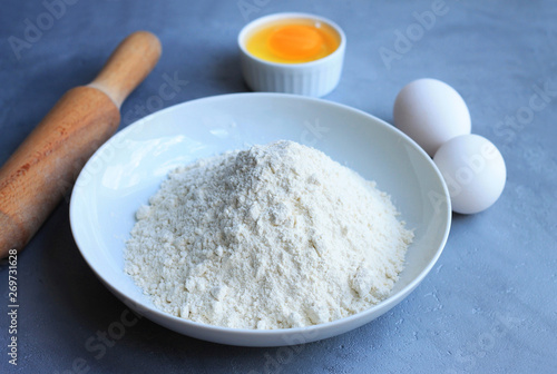 Plate with flour, eggs and a rolling pin on a gray background.